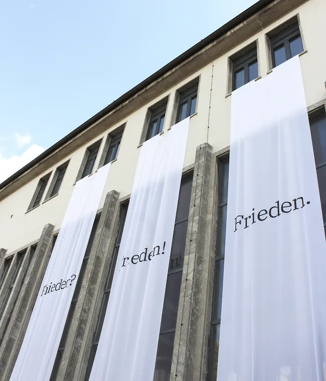 Weiße Banner mit den Worten ‚Frieden?‘, ‚Frieden!‘ und ‚Frieden.‘ hängen an der Fassade eines Gebäudes mit mehreren Fenstern.